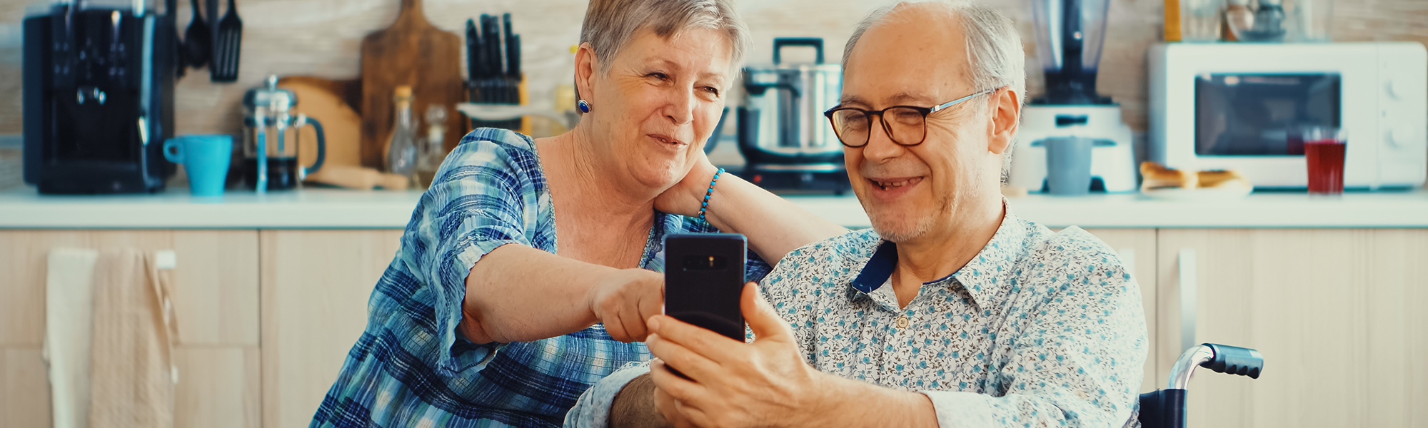 Lächelnde Senioren und ihr Ehemann im Rollstuhl nutzen Smartphone in der Küche